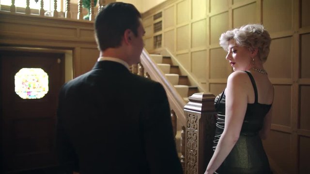 Attractive blonde woman wearing a classic vintage fifties dress waits at the bottom of a staircase.  She turns and a handsome man in a suit walks into frame. Originally recorded in 4K.