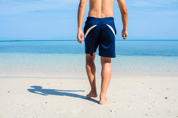 Man with no shirt walking barefoot on white sand beach into the