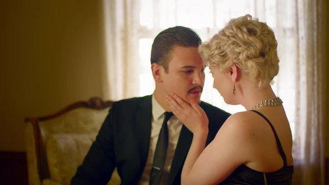 Attractive woman wearing a classic vintage fifties dress and sitting on a chaise lounge pulls handsome man towards her before camera pans away.  Recorded in 4K and tinted.