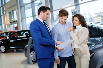 Salesman talking to a young couple at the dealership showroom.