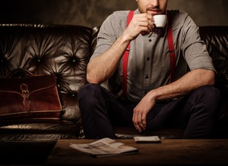 Young handsome old-fashioned bearded man with cup of coffee sitting on comfortable leather sofa on...
