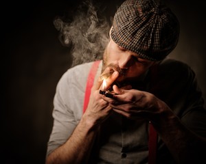 Serious old-fashioned man in tweed hat lighting a cigar on dark background.