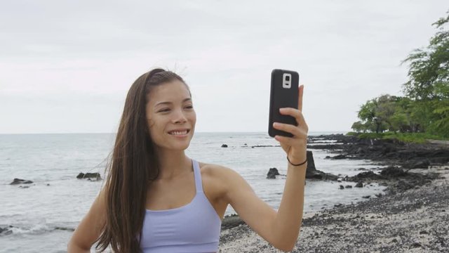 Phone app - Fitness woman looking at smartphone on beach. Running woman using smartphone application resting relaxing with water bottle after workout exercise on beach outside. Mixed race Asian girl. 