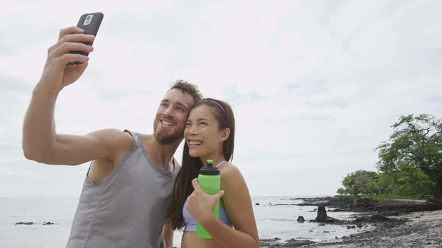 Fitness couple taking selfie after running workout. Good looking young man and woman taking a self-portrait picture with smart phone camera after exercising on a beach during summer vacation travel.