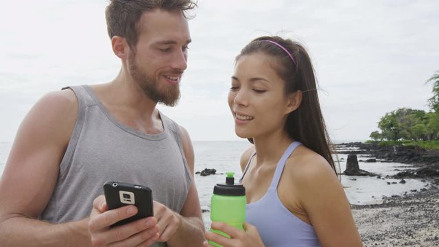 Friends looking at phone app after running workout fitness. Good looking young man and woman using smartphone relaxing after exercising on a beach. Mixed race couple. RED EPIC.