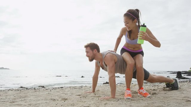 Fitness couple laughing having fun together doing funny push-up on beach during workout. Woman playful h sitting on boyfriend to test his strength with heavy weight. RED EPIC.