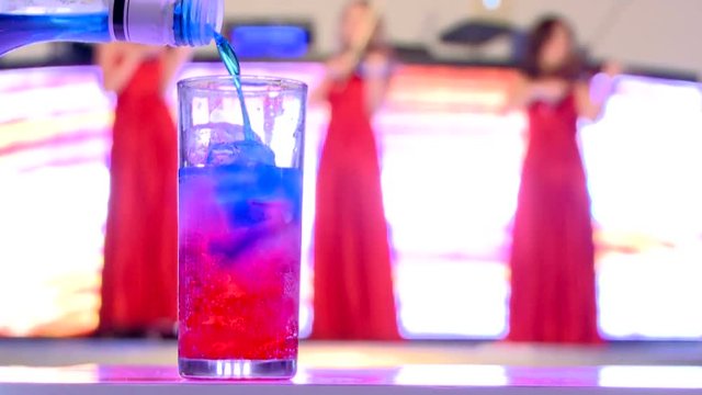 Night party. Mix cocktail. blue and red drinks mixing. glass with ice from the bottle. with 3 women in red dress playing violin on the stage in the background at the party, family celebration.