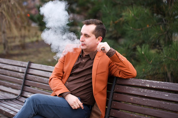 A young man sits on a bench and smokes an electronic cigarette.