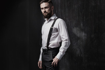 Attractive bearded man in a white shirt and suspenders standing near dark wall. He is strong, courageous and serious. Dark room, night and harsh shadows.