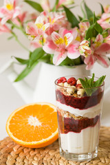 Homemade parfait serverd in high glasses with yogurt and organic raspberries, nuts and mint. Delicious snack for healthy life.  Breakfast on a straw tablemat with spoons and flowers and half of orange