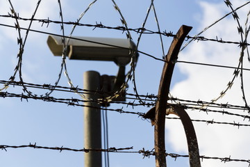 Security camera behind barbed wire fence stretched around prison walls