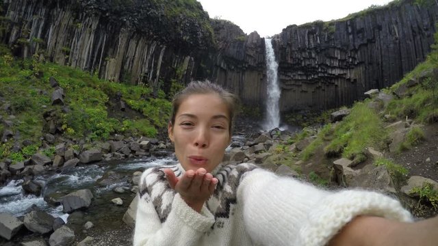 Happy woman blowing kiss against majestic Svartifoss waterfall. Female is visiting famous tourist attraction of Iceland. Skaftafell, Vatnajokull National Park, Iceland. ACTION CAMERA.