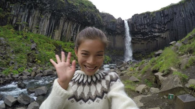 Smiling beautiful woman waving against majestic Svartifoss waterfall. Female is visiting famous tourist attraction of Iceland during vacation. Svartifoss waterfall, Skaftafell. ACTION CAMERA.