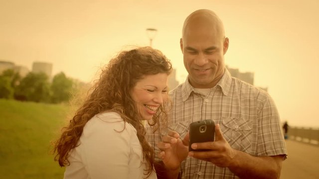 Warm tinted clip of laughing and smiling couple use a smart phone with Detroit city skyline visible in background.  Slow motion, recorded hand-held at 60fps in 4K.