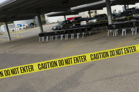 Yellow do not enter caution tape stretched across a parking lot with tables and chairs set for a company picnic. 