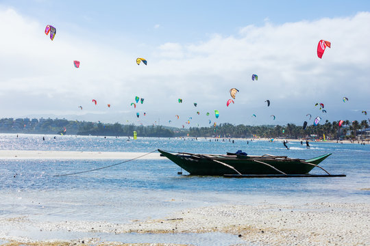 Kitesurfers enjoying wind power on Bulabog beach, Boracay island, Philippines. Empty boat moored in foreground.
