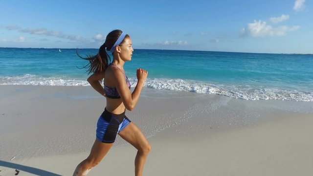 Woman jogging and running on beach. Fit young female runner in sportswear is training on idyllic beach. Runner is exercising in nature on sunny day. ACTION CAMERA TRACKING SHOT.