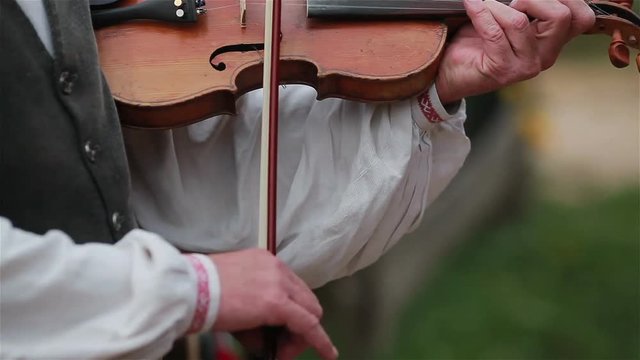 Man in a white traditional shirt playing violin outdoors in street with unfocused musicians and audience on the background. Close up