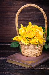 Fototapeta na wymiar Yellow roses in a basket, vintage book on wooden background. Free space for your text.