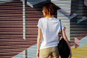 young girl with a backpack wearing a white blank t-shirt standing on a graffiti wall background.back view