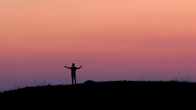 A young man (silhouette) is standing on top of a hill and raises his arms embracing the great view and a feeling of freedom, accomplishment and success. Filmed in 4k UHD high resolution.
