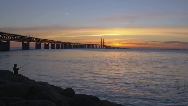 Oresundsbron at sunset. The bridge between Sweden and Denmark. Cars drive over the bridge and underneath the bridge goes a train. A fisherman stands fishing