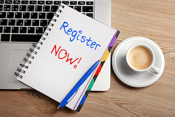 Register now written in notebook, laptop and cup of coffee on table, top view