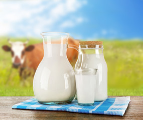 Pitcher, jar and glass of milk on wooden table against cow and blue sky background