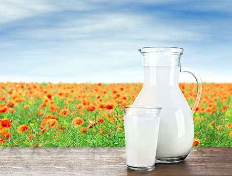 Pitcher, glass of milk on wooden table against poppies and blue sky background