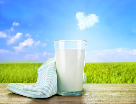 Glass of milk on wooden table against green field and blue sky background
