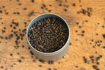Still life with black peppercorn inside round silver jar and scattered on vintage wooden background. Horizontal photo closeup