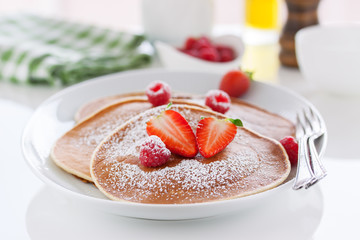 Homemade buttermilk american pancakes with fresh strawberry and raspberry on a white plate on a white table for breakfast, closeup, selective focus..