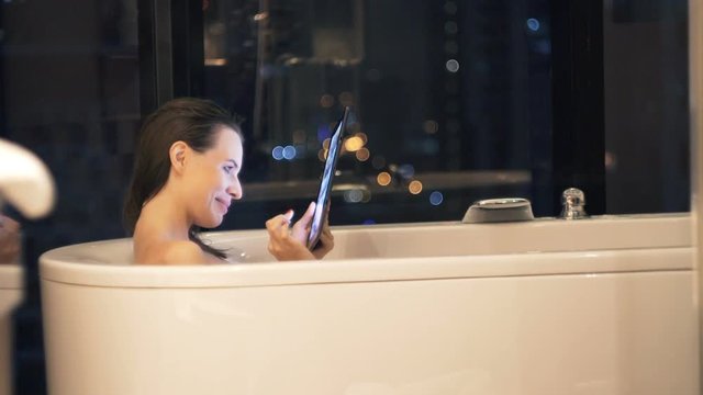 Young, pretty woman browsing photos on tablet during bath at night
