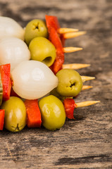 Vegetable skewers of olives, capiscum, pickles and baby onions lying on grey stone surface as seen from above