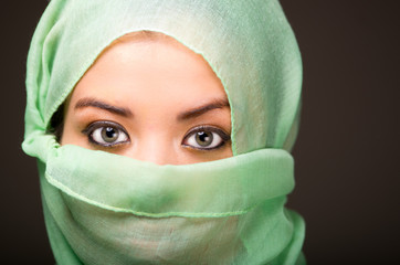 Woman wearing turquoise scarf covering face only revealing beautiful green eyes, grey dark background