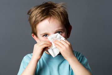 healthcare learning - smiling young preschooler with red hair and blue eyes using a tissue after a cold or having rhinitis allergies, grey background studio.