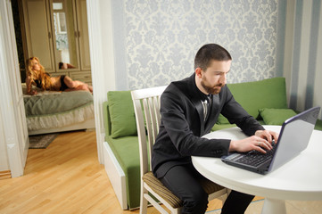 Young man in black suit and a tie is sitting at the table using notebook. Sexy beautiful woman in black lingerie is laying on the bad on the background. Relationship lifestyle concept