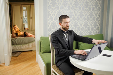 Young husband in black suit and a tie is sitting at the table using notebook. Sexy beautiful wife in black lingerie is laying on the bed on the background. Relationship lifestyle concept