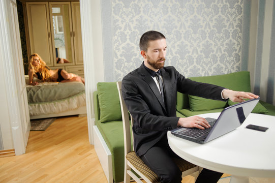 Young man in black suit and a tie is sitting at the table using notebook. Sexy beautiful lady in black lingerie is laying on the bed on the background. Relationship lifestyle concept