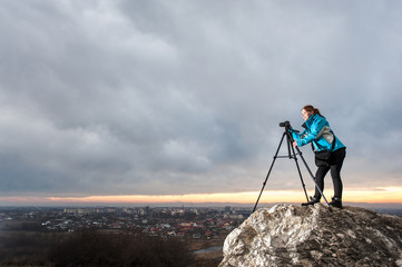 Young female photographer is standing with her camera on tripod on the big rock at city overview point. Girl taking picture of the cityscape in the evening. Rain clouds and sunset in the background