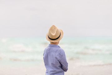 Fototapeta na wymiar Portrait of adorable kid boy in straw hat and sun glasses walking on ocean beach and looking at the waves. Vacations by the sea. Outdoor.