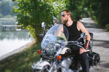 Fototapeta na wymiar Handsome biker in sunglasses with long hair and beard sitting on his motorcycle near the lake on a sunny day. Tilt shift lens blur effect.