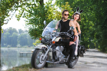 Fototapeta na wymiar Sexy couple of bikers on the motorcycle near the lake. Lovely woman in rabbit costume, handsome man with beard wearing sunglasses. Hot outdoors summer day. Tilt shift lens blur effect