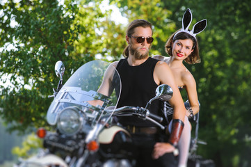 Obraz premium Portrait of sexy couple of bikers on the motorcycle near the lake. Girl in bunny costume, brutal man with beard wearing sunglasses. Hot outdoors summer day. Tilt shift lens blur effect