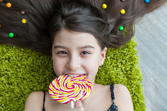 Little girl with dark hair lying on the floor among the sweets. Top view of a big yellow and pink lollipops in hands. Many candies in her hair. smile, joy and emotion on the face of the child.