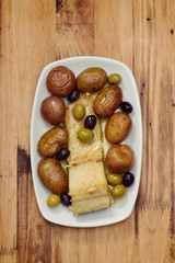 fish with potato and olives on white dish on brown wooden background