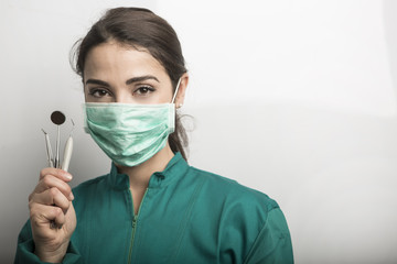 Primo piano di odontoiatra donna con mascherina, camice Verde che ci mostra i suoi  attrezzi da lavoro -  sfondo grigio 