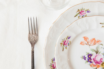 Two white plates a fork on white background. White porcelain dishes with colored flowers and golden edge on white tabecloth. Wedding table setting. Festive table setting.