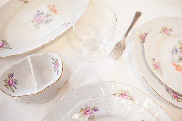 White plates a fork a wineglass on white background. White porcelain dishes with colored flowers and golden edge on white tabecloth. Wedding table setting. Festive table setting.