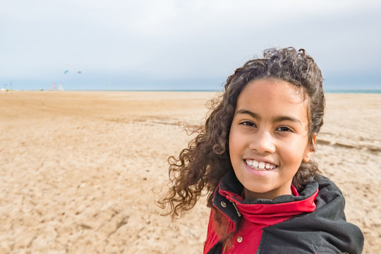 Adorable young girl wearing red coat, walking on beach in Spain in winter. Beautiful brazilian child with long curly hair enjoying wind and sun outside, image for children concept family blog. 